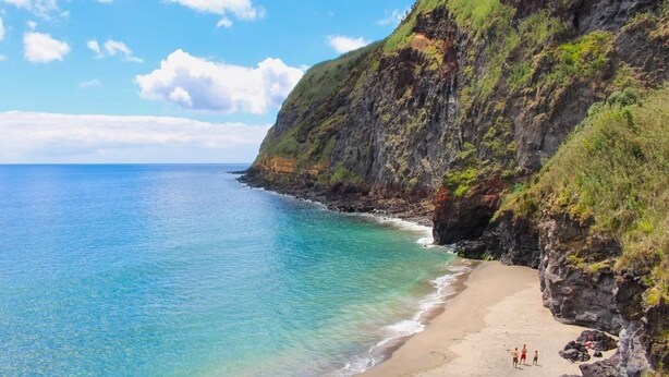 A coastal beach with rocky cliffs and clear blue water.