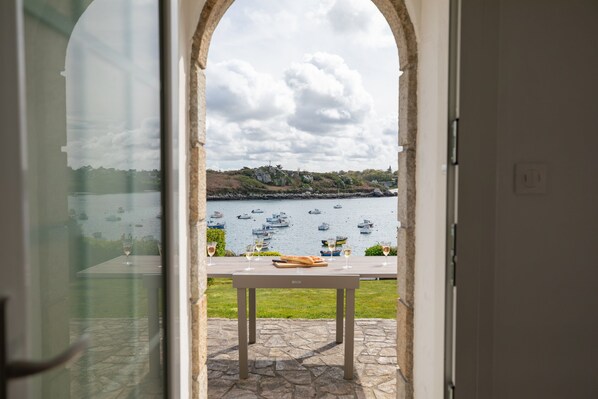 Vista a través de un arco hacia un patio con mesa y sillas, con vista a un cuerpo de agua y barcos.