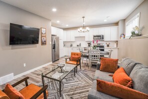A modern kitchen with white cabinets, a dining area with a glass table, and a living room with a gray sofa and orange cushions. - Modern Comfort in Westgate (Grande Prairie)