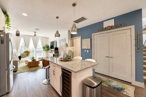 A modern kitchen with a central island, pendant lighting, and a view into a living area with a sofa and chairs. - Bentley Sky Lounge with Downtown View (Dallas)