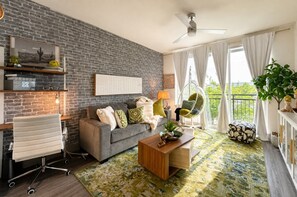 A living room with a brick wall, a grey sofa, a wooden coffee table, and a view of greenery through large windows. - Bentley Sky Lounge with Downtown View (Dallas)