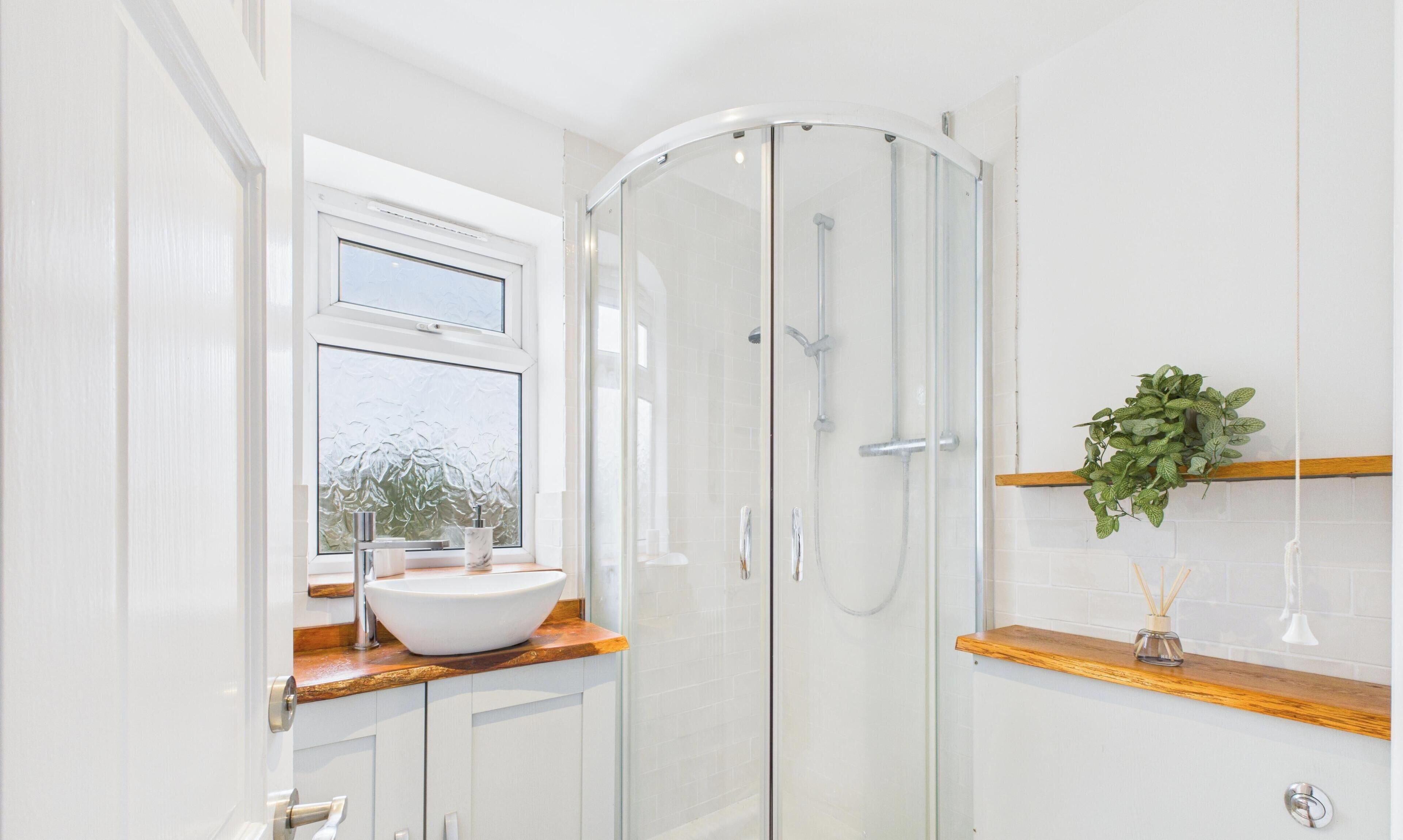 Une salle de bain moderne avec une douche à l’italienne en verre, un lavabo blanc doté d’un plan de toilette en bois et une fenêtre en verre dépoli.