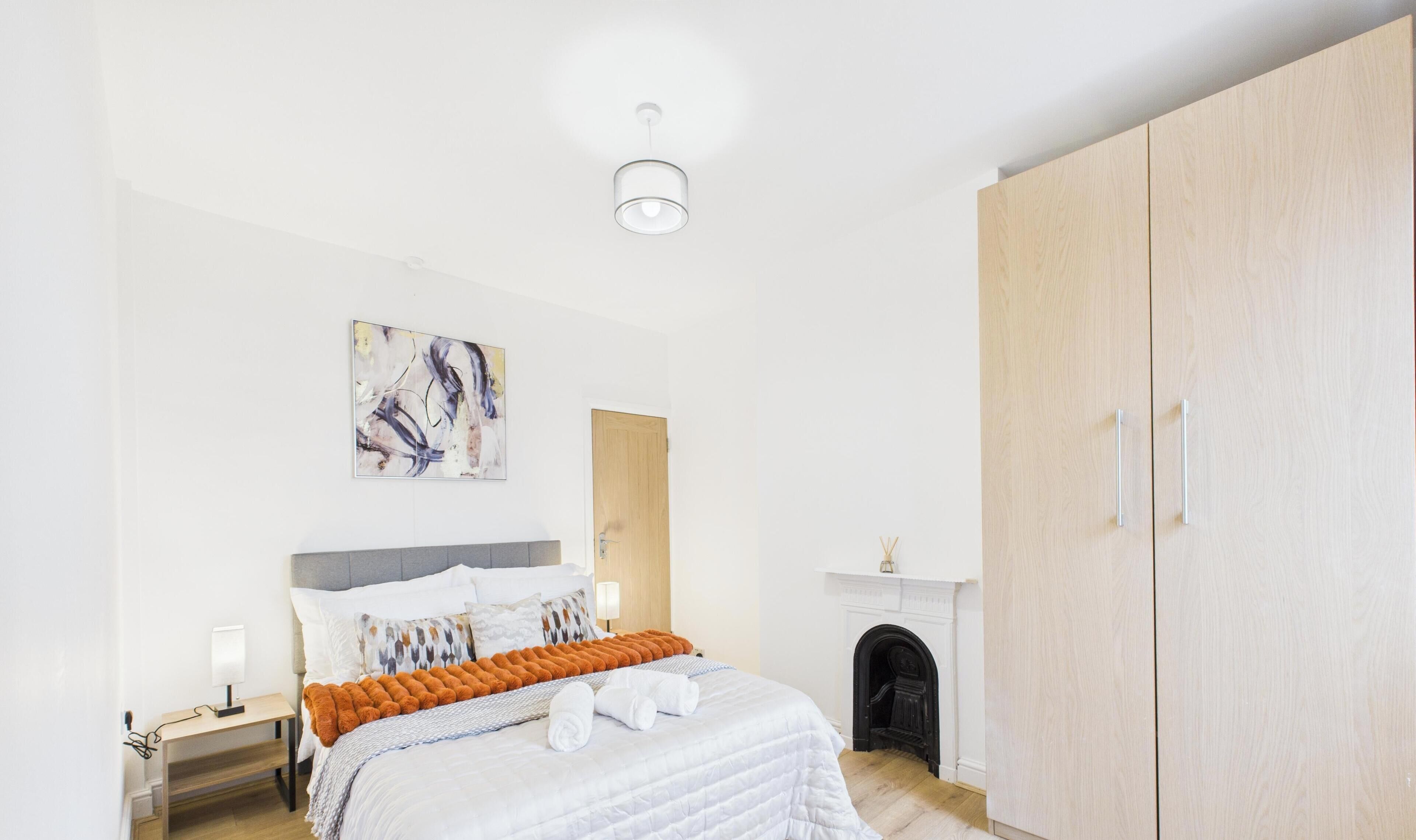 A bedroom with a bed, a nightstand, a wall-mounted lamp, a painting, a wooden wardrobe, and a fireplace.