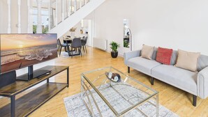 En rummelig stue med trappe, et fladskærms-tv, en grå sofa og et glasbord.
