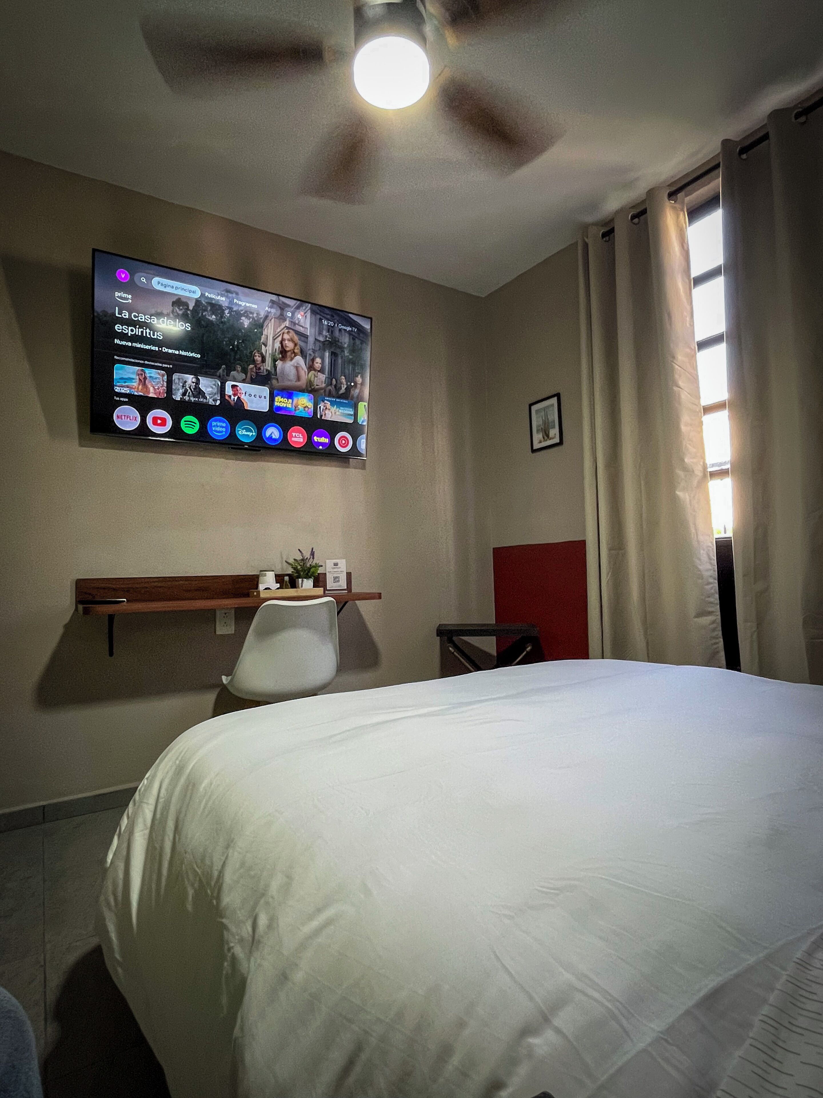 A hotel room with a bed, a desk, a chair, a television mounted on the wall, and a ceiling fan.