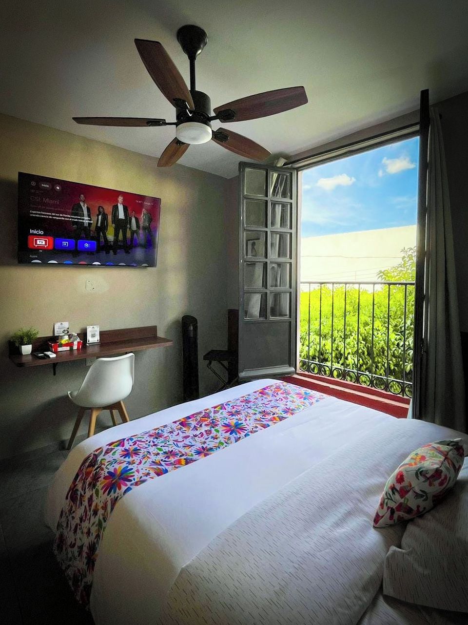 A hotel room with a bed, a ceiling fan, a TV on the wall, a balcony with a view, and a desk with a chair.