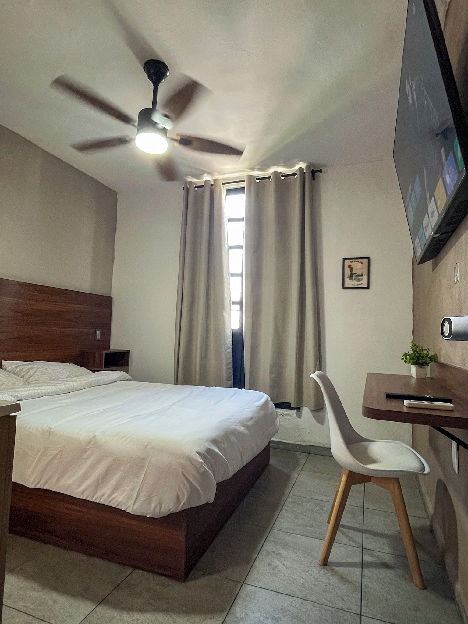 Habitación de hotel con cama, escritorio, silla, ventilador de techo y televisión en la pared.