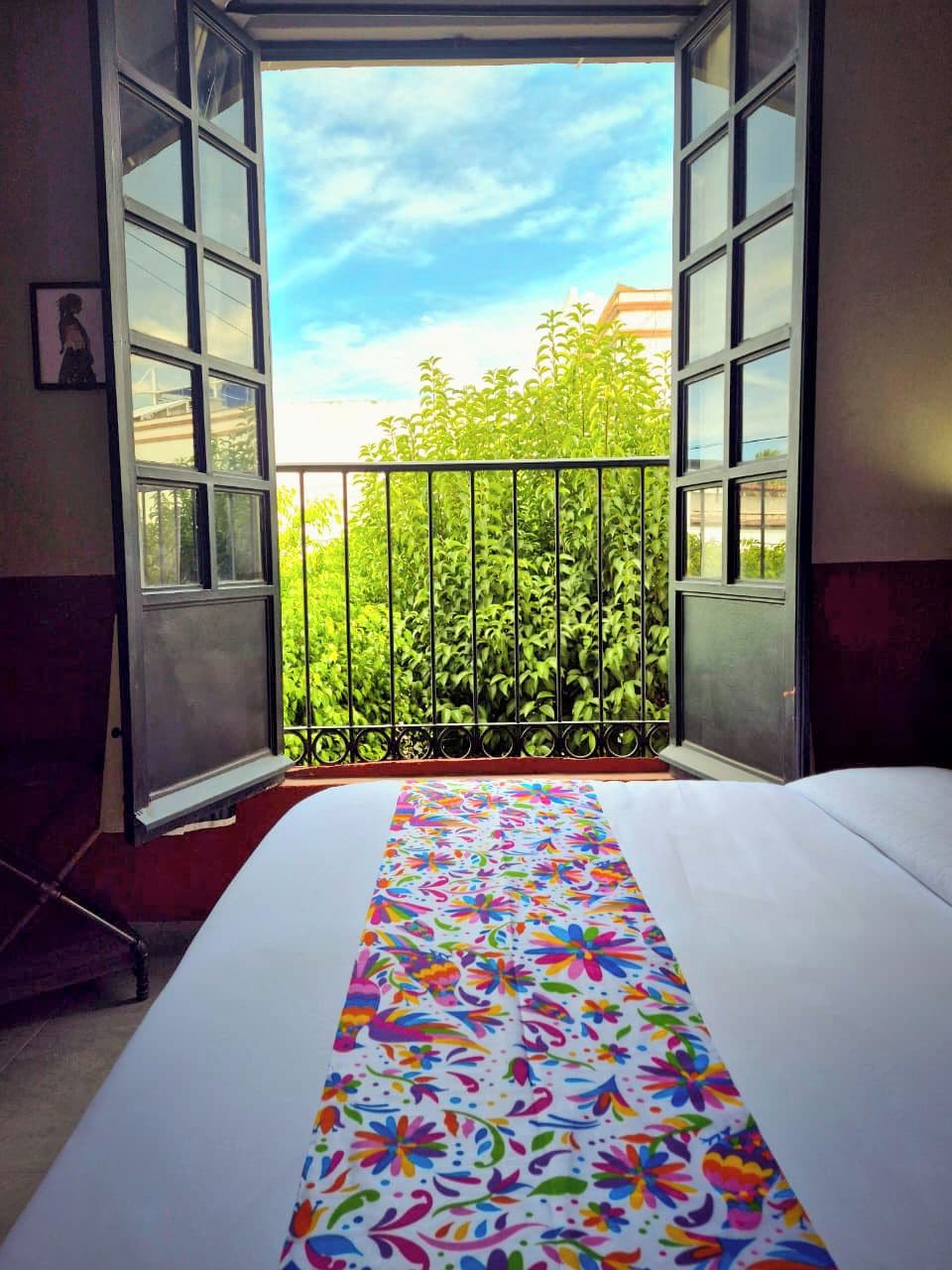 Una habitación con una cama, un ventanal con vistas abiertas a un balcón y una colcha de colores.