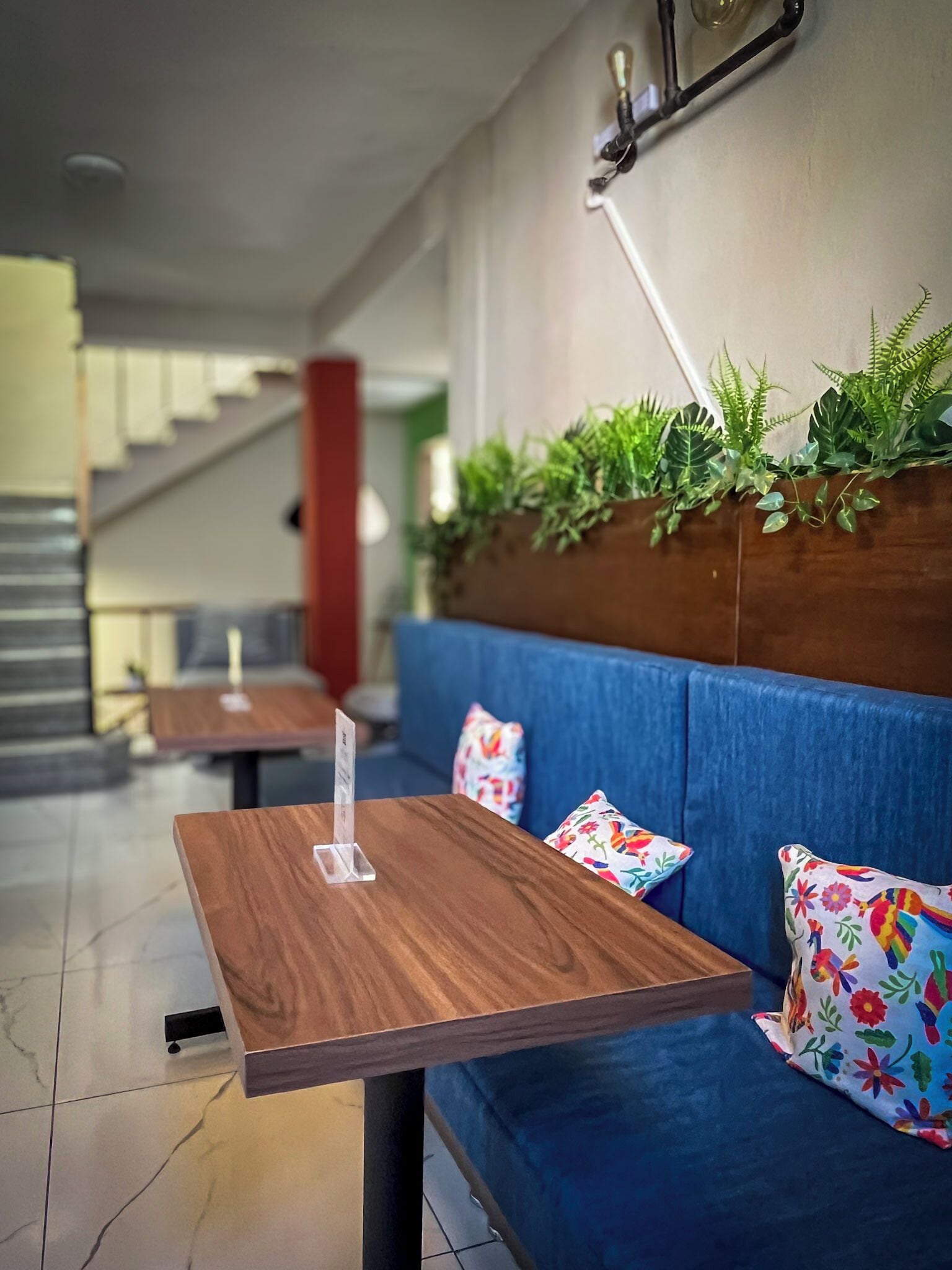 Un área de descanso con una mesa de madera y un sofá azul, una escalera al fondo y plantas verdes en la pared.