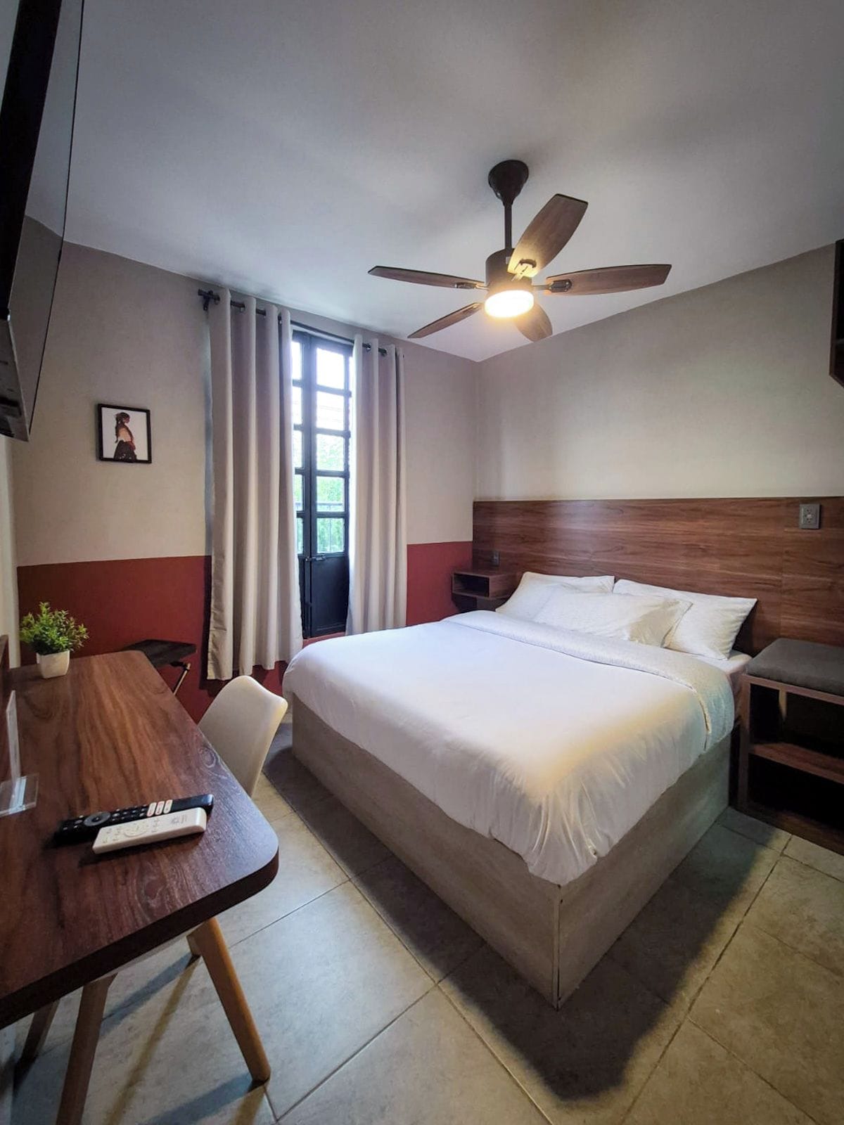 Una habitación de hotel con cama, escritorio, silla, ventilador de techo y ventana con cortinas.