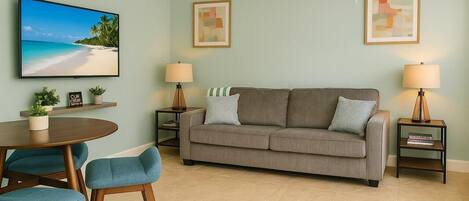 Una sala de estar con un sofá, dos mesas auxiliares con lámparas, un televisor en la pared, un ventilador de techo y una zona de comedor con sillas y una mesa.