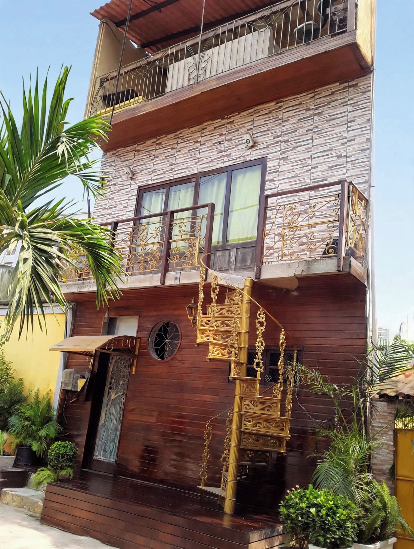 Un edificio de varios pisos con fachada de madera, un balcón con barandillas de metal ornamentadas y un pequeño jardín con plantas en macetas.
