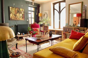 A room with a sofa, a table with a bowl, a bookshelf, a radiator, and a painting on the wall. - A garden in the city... (Nantes)