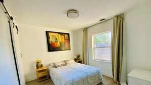 A bedroom with a bed, a painting, a window with curtains, a bedside table with a lamp, and a nightstand with flowers. - Cherry red door cottage with AC in cool Phoenix C (Phoenix)