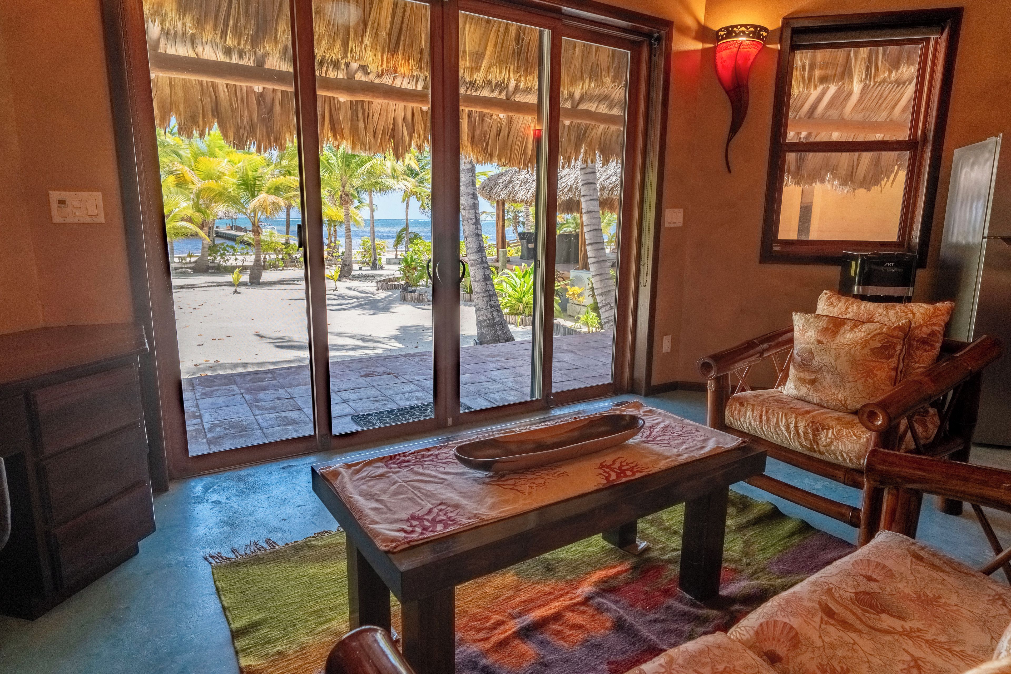 A room with a large sliding glass door leading to a beach view, a wooden table with a bowl, a cushioned chair, and a wall-mounted light fixture.