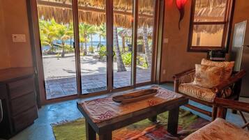 A room with a large sliding glass door leading to a beach view, a wooden table with a bowl, a cushioned chair, and a wall-mounted light fixture.