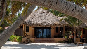 A thatched-roof structure with a sandy ground, palm trees, and a bench. - Casa Pisces (San Pedro)