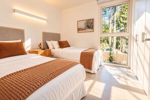 Un dormitorio con dos camas, una mesita de noche, un cuadro enmarcado y una ventana con vista a árboles.