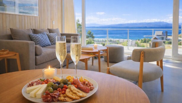 A dining area with a view of the sea, a plate of food, two glasses of champagne, and a lit candle. - Signature Lake View Luxury Residences A01 (San Carlos de Bariloche)