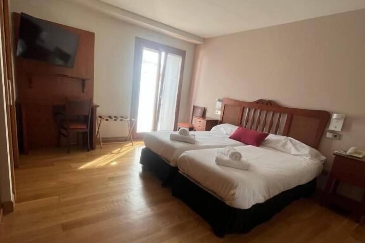 A hotel room with a wooden floor, a bed with white bedding, a television mounted on the wall, and a sliding door leading to a balcony.