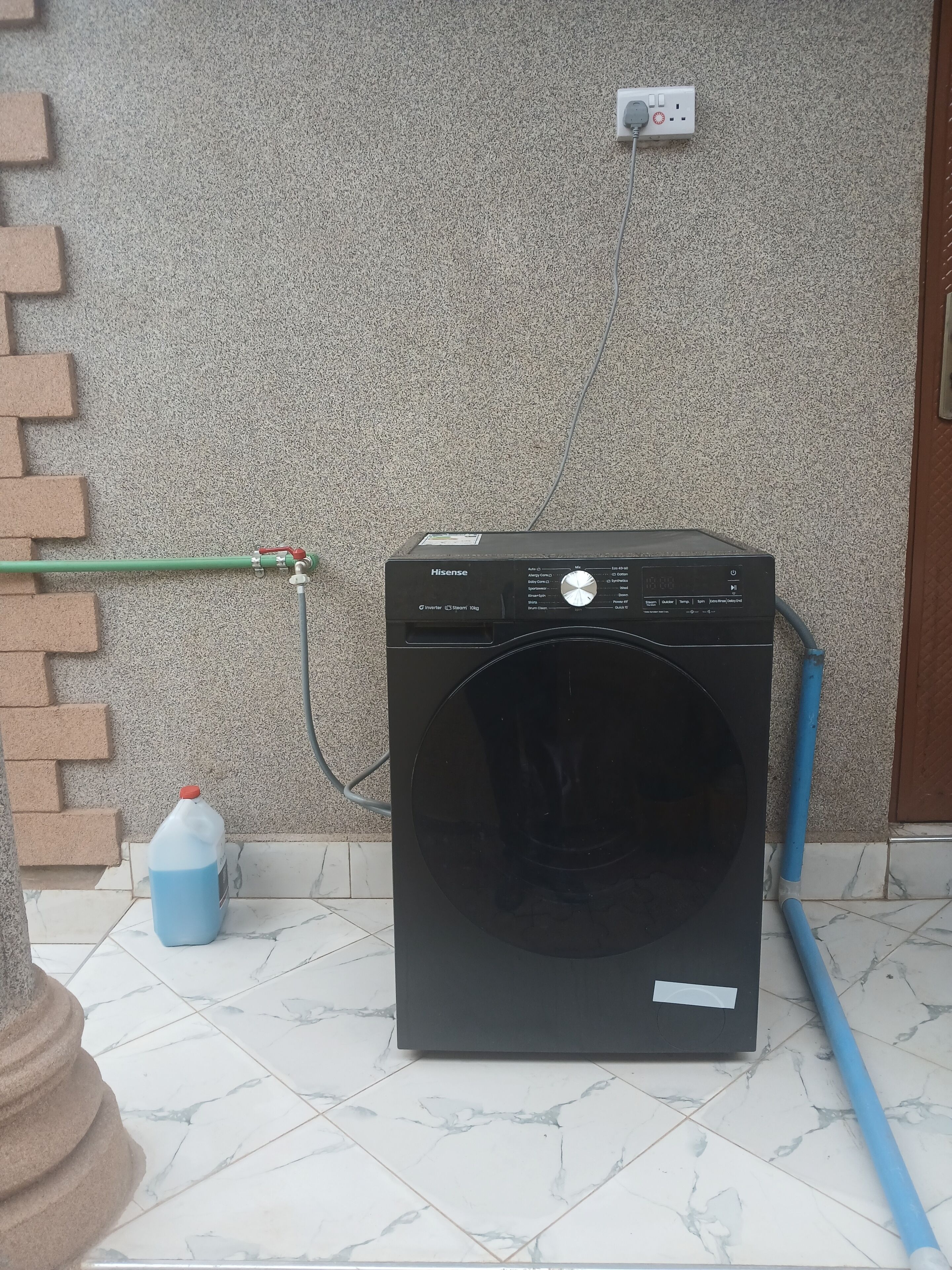 Una lavadora Hisense inverter con vapor de 1100g de color negro, con una manguera verde y otra azul conectadas, ubicada sobre un suelo de baldosas frente a una pared con textura.