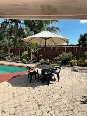 Un espace patio avec une piscine, une table entourée de chaises et un parasol.
