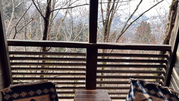 Un banco de madera con una mesa pequeña, dos sillas estampadas y una barandilla de madera que ofrece vistas a árboles y montañas.