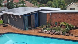 A swimming pool with a tiled deck, a small building with a dark facade and blue accents, and a garden area with various plants and rocks. - Quaint cottage in Glenwood, Durban (Durban)