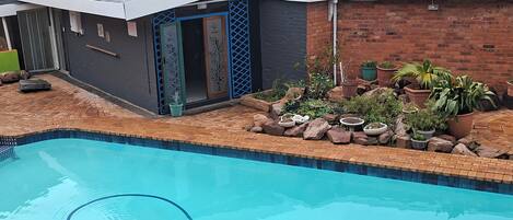 A swimming pool with a tiled deck, a small building with a dark facade and blue accents, and a garden area with various plants and rocks.