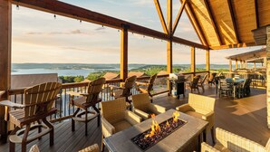 A covered outdoor deck with wooden furniture, a fire feature, and a view of a lake.