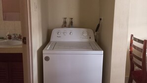 A compact, white Whirlpool washer in a small laundry room with a tiled floor, a wooden chair, and a sink with a pink mat. - Fabulous 1-bedroom apartment with AC in Saint Michael (Saint Michael)