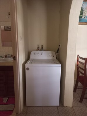 A compact, white Whirlpool washer in a small laundry room with a tiled floor, a wooden chair, and a sink with a pink mat. - Fabulous 1-bedroom apartment with AC in Saint Michael (Saint Michael)