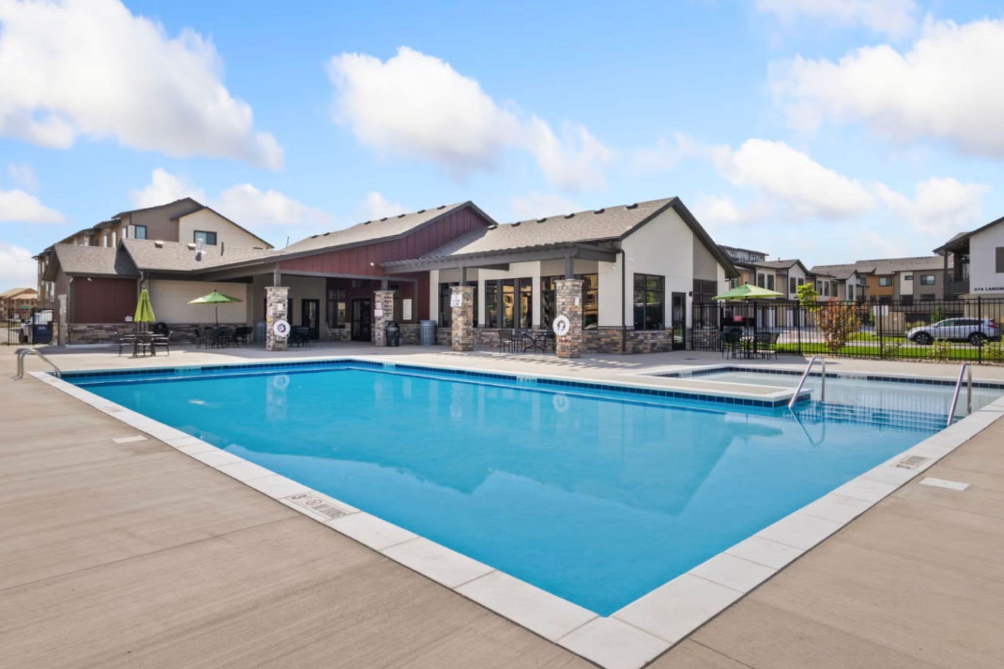 A rectangular swimming pool with a tiled deck surrounding it, adjacent to a building with outdoor seating and a covered patio area.
