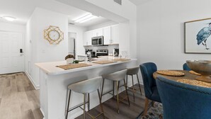 Et moderne køkken med hvide skabe, en central ø med barstole og et spiseområde med en blå stol og en stor skål.