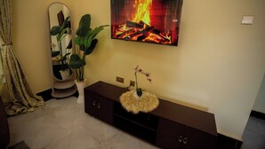 Un salon avec un téléviseur fixé au mur qui diffuse une image de feu de cheminée, une plante en pot, un miroir et un meuble sombre avec des tiroirs.
