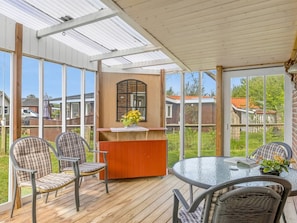 Eine überdachte Terrasse mit einem runden Glastisch, vier Stühlen, einer Holzbank und einem kleinen Tisch mit Blumen.