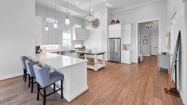 A modern kitchen with a central island, bar stools, pendant lighting, and a view into the dining area. - Luxury Beachfront Ocean Views Pool Access Sleeps 15 (Bolivar Peninsula)