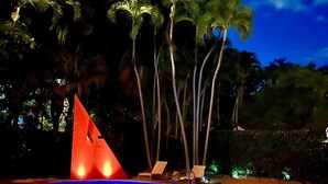 Een zwembad met een geometrisch sculptuur, omringd door palmbomen en ligbedden.