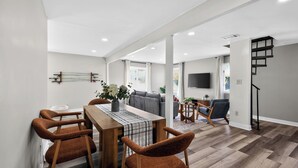 A modern living room with a dining area, a staircase, and a flat-screen TV mounted on the wall. - Infrared Sauna, Firepit, Kayaks, Walk 2 Launch, Authentic WI Lake Life (Poynette)
