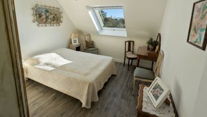 Une chambre avec un plafond en pente, un puits de lumière, un lit recouvert d’une couverture blanche, un bureau avec une chaise et une petite table sur laquelle est posée une plante.