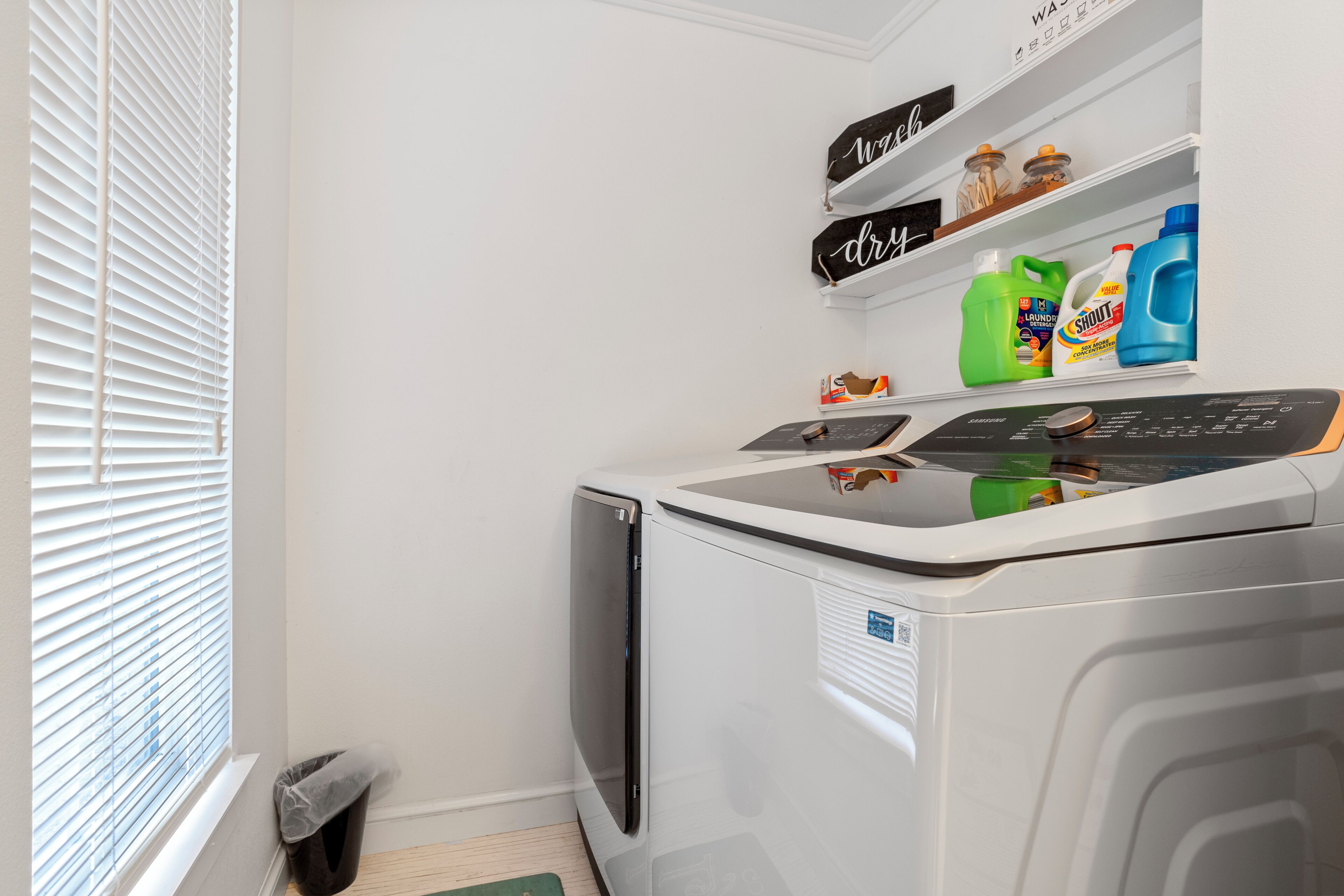 A laundry room with a washing machine, shelves with cleaning supplies, a trash bin, and a window with blinds.