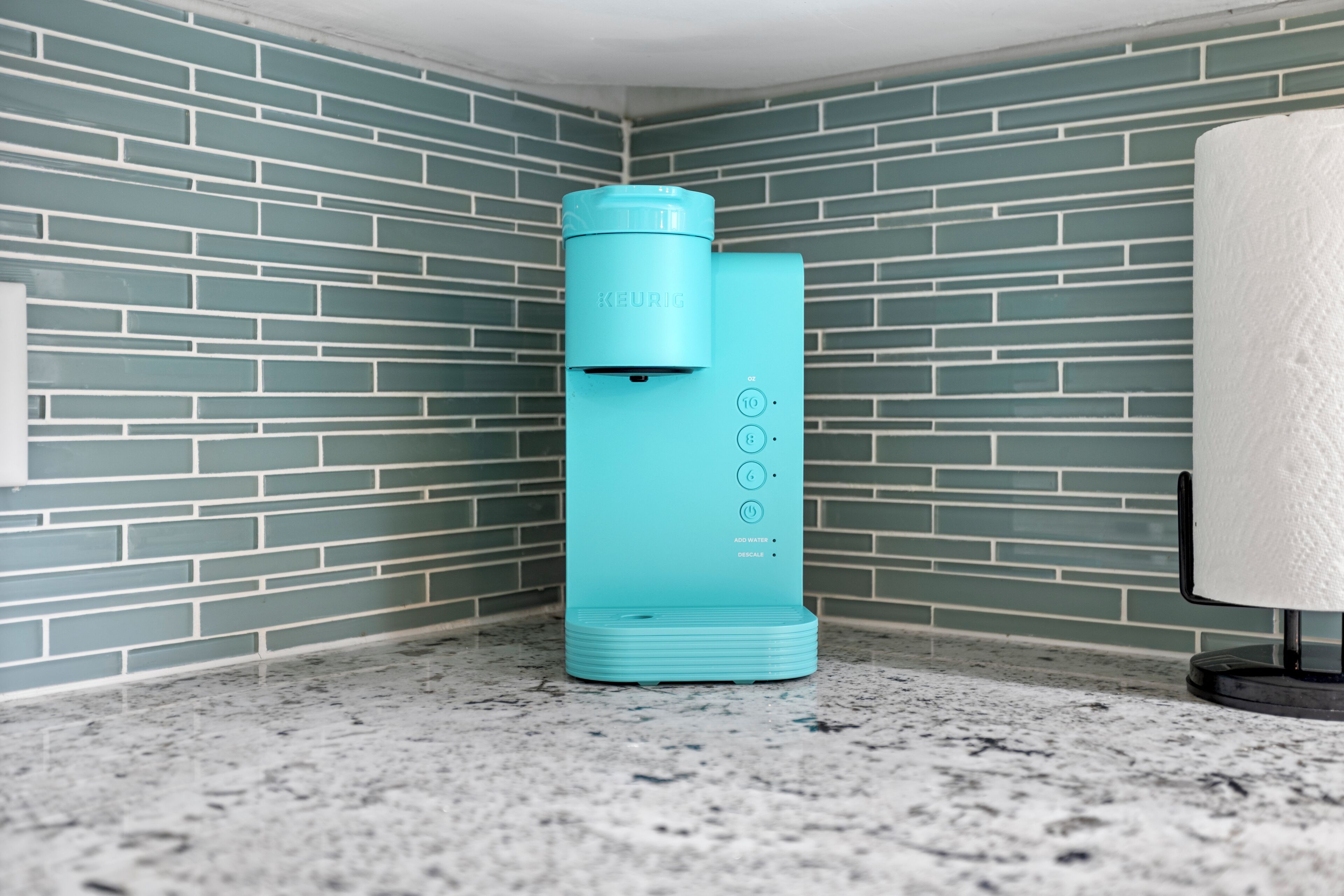 Una cafetera color turquesa con depósito de agua, un dispensador de toallas de papel y un salpicadero de azulejos en una esquina.