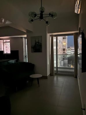 Ein Wohnzimmer mit einer Couch, einem kleinen Tisch, einer Chandelieber, einem Bild an der Wand und einer Glastür, die auf einen Balkon mit Stadtblick führt.