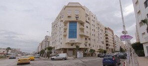 Ein mehrstöckiges Hotel mit einem zentralen, blauen Eingang, zahlreichen Balkonen und einem Parkplatz mit Autos.