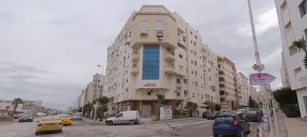 A multi-story hotel with a central blue entrance, multiple balconies, and a parking area with cars. - Luxury & Fully Equipped | Safe & Terrace | All Amenities within Walking Distance (Tunis)