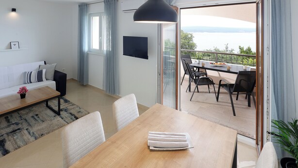 Una sala de estar moderna con sofá, mesa de centro y mesa de comedor. Cuenta con un ventanal con cortinas, un televisor montado en la pared y un balcón con mobiliario exterior.