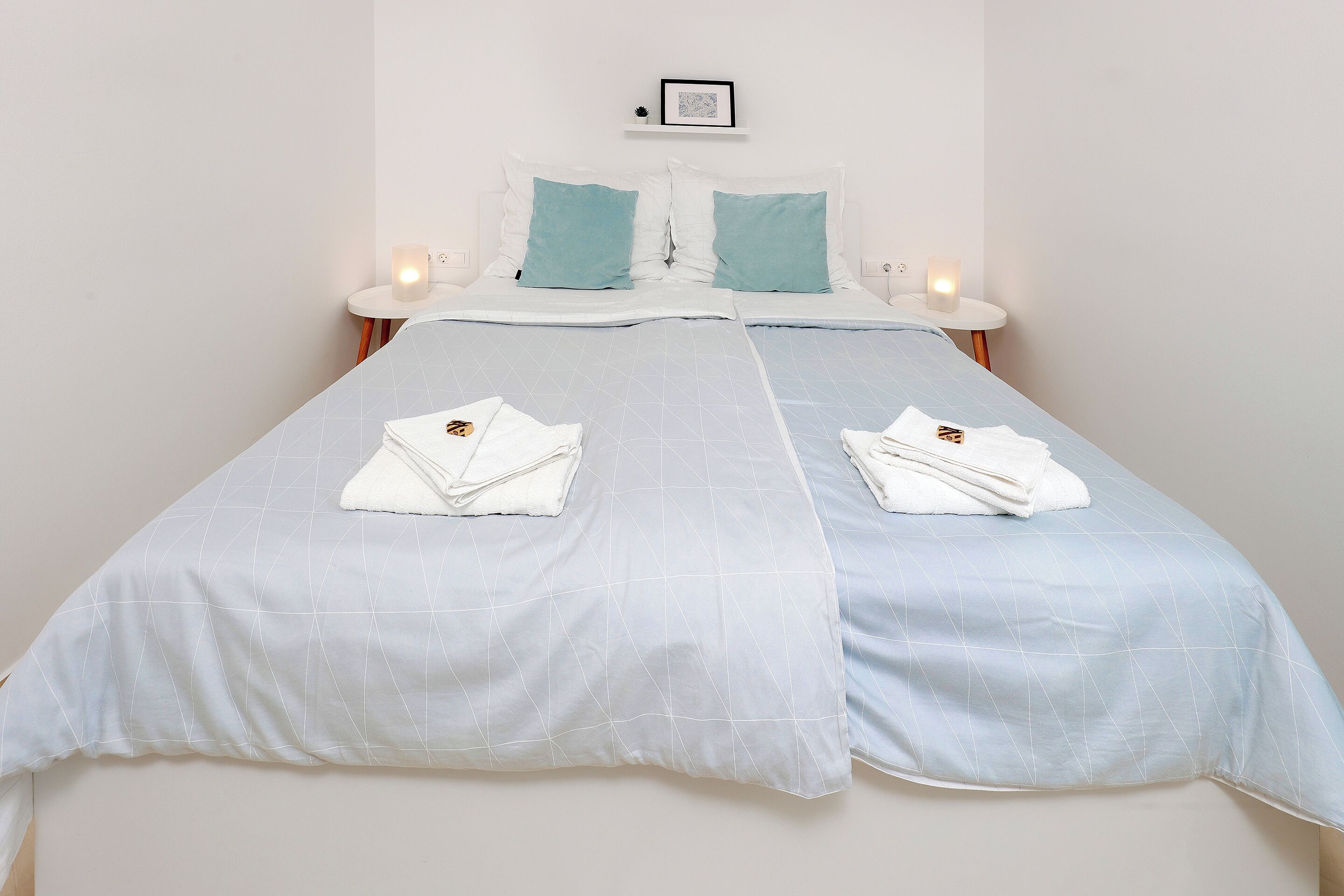 A double bed with white and light blue bedding, two bedside tables with candles, and a framed picture on the wall.