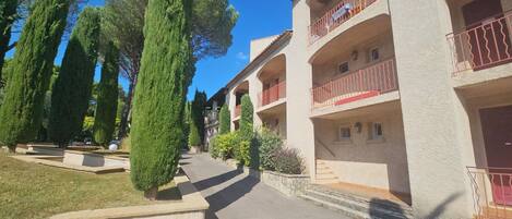 A multi-story building with balconies, a paved pathway, and neatly trimmed trees.