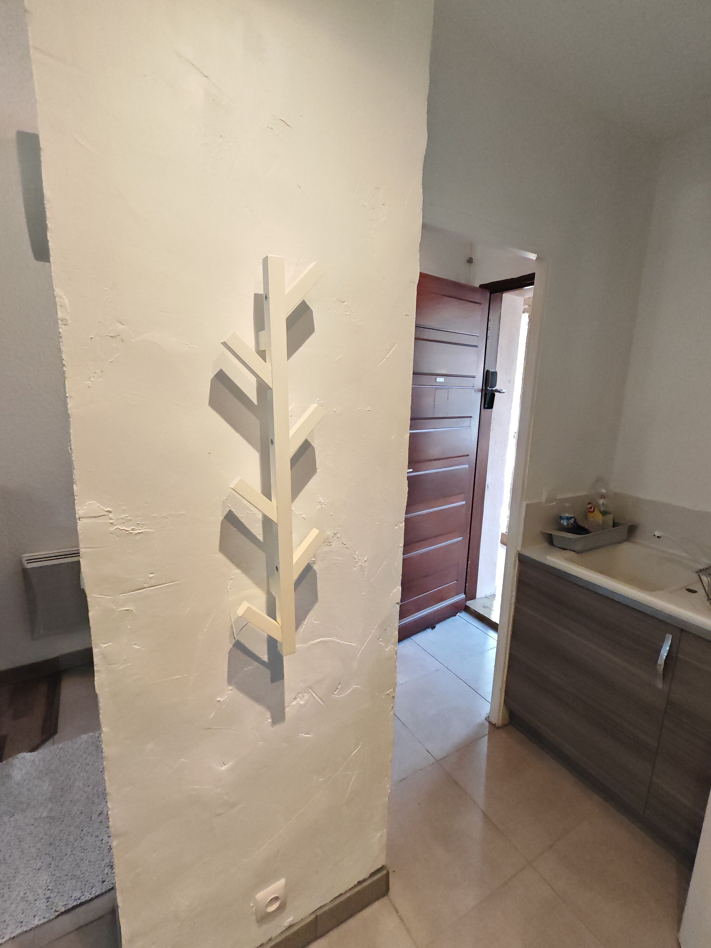 Une pièce avec un mur blanc, une porte en bois, un coin cuisine avec un plan de travail et des placards, et un sol carrelé.