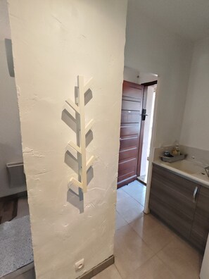 Ein Zimmer mit weißer Wand, einer Holztür, einer Küchenzeile inklusive Arbeitsplatte und Schränken sowie einem Fliesenboden.